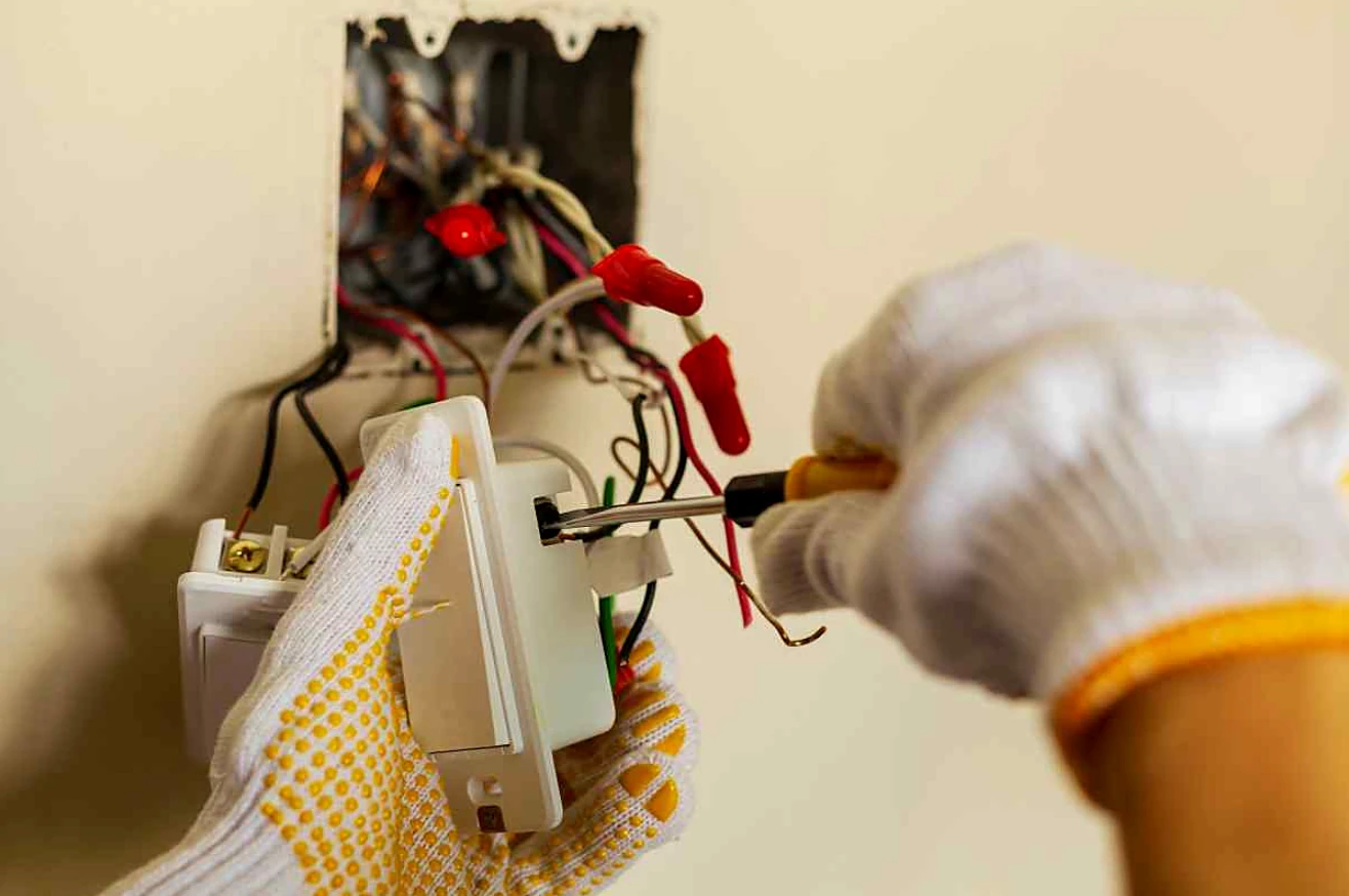 An electrician in work gloves rewiring a damaged switch box.